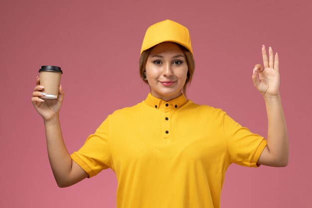 Вид спереди женщина-курьер в желтой форме с желтым плащом держит кофе с улыбкой на розовом фоне.