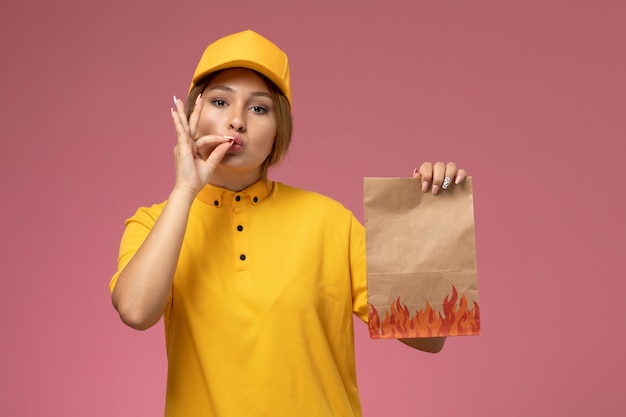 Corriere femminile di vista frontale in capo giallo uniforme giallo che tiene il pacchetto di consegna sul lavoro di consegna uniforme sfondo rosa