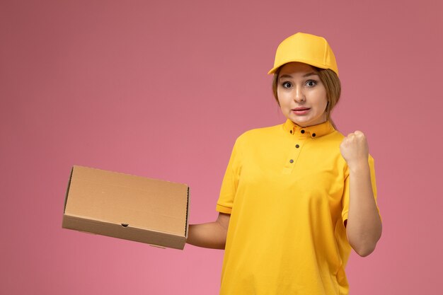 Front view female courier in yellow uniform yellow cape holding food box on pink desk uniform delivery female color