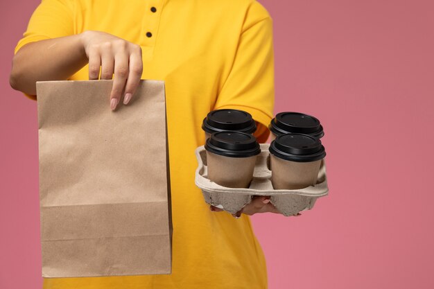 Вид спереди женщина-курьер в желтой униформе с желтым плащом держит пакет с едой и кофе на розовом фоне.
