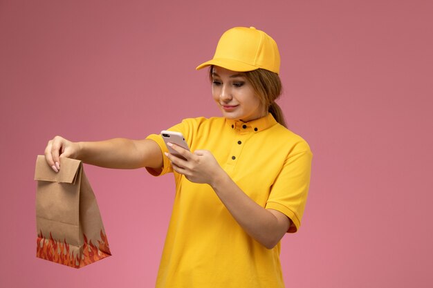 ピンクの背景に食品パッケージと電話を保持している黄色の制服黄色のケープの正面図女性宅配便制服配達作業色の仕事