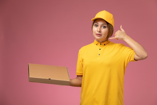 Front view female courier in yellow uniform yellow cape holding food package showing phone call gesture on the pink desk uniform delivery work color