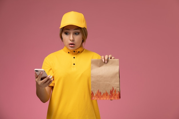 Front view female courier in yellow uniform yellow cape holding food package and using smartphone on pink background uniform delivery work color job