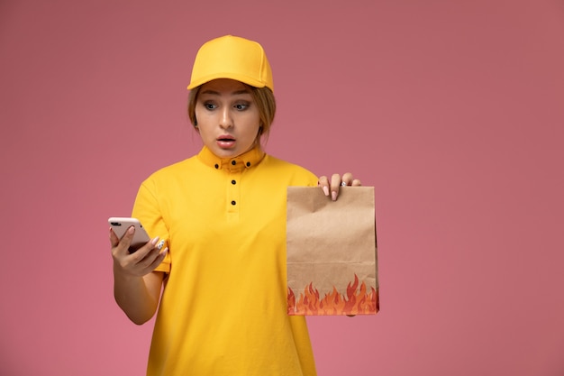 음식 패키지를 들고 분홍색 배경에 스마트 폰을 사용하여 노란색 유니폼 노란색 케이프의 전면보기 여성 택배 유니폼 배달 작업 색상 작업