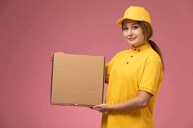 Front view female courier in yellow uniform yellow cape holding food package with slight smile on pink desk uniform delivery female work color