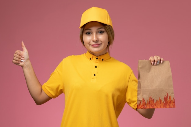 Corriere femminile di vista frontale in mantello giallo uniforme giallo che tiene il pacchetto alimentare con il sorriso sul lavoro di colore di lavoro di consegna uniforme sfondo rosa