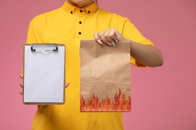 Front view female courier in yellow uniform yellow cape holding notepad and food package on pink background uniform delivery work color job