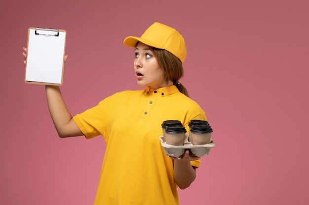 Front view female courier in yellow uniform yellow cape holding notepad plastic coffee cups on pink background uniform delivery work color job