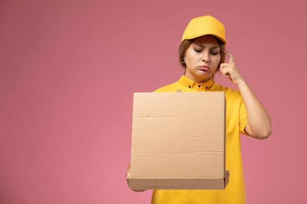 Front view female courier in yellow uniform yellow cape holding and opening food box on the pink desk uniform delivery female color