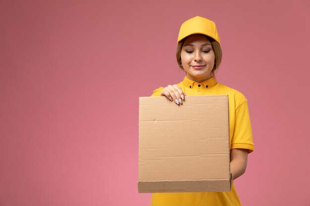 Front view female courier in yellow uniform yellow cape holding and opening food box on the pink desk uniform delivery female color