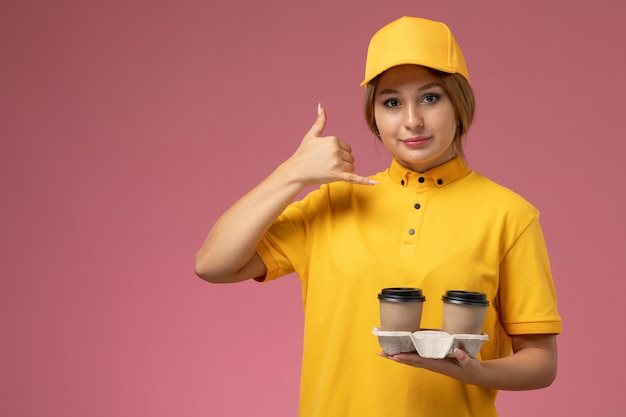 ピンクの机の上のプラスチック製の茶色のコーヒーカップを保持している黄色の制服黄色のケープの正面図女性宅配便制服配達女性