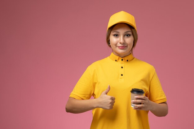 ピンクの机の上に笑顔でプラスチック製の茶色のコーヒーカップを保持している黄色の制服黄色のケープの正面図女性宅配便制服配達女性の色