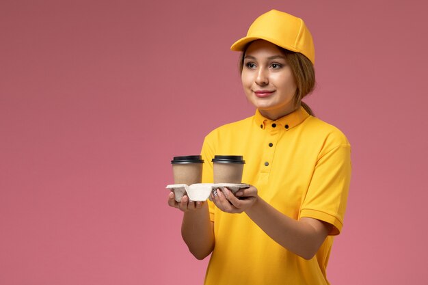 플라스틱 갈색 커피 컵을 들고 분홍색 책상 유니폼 배달 여성 색상에 전달하는 노란색 유니폼 노란색 케이프의 전면보기 여성 택배