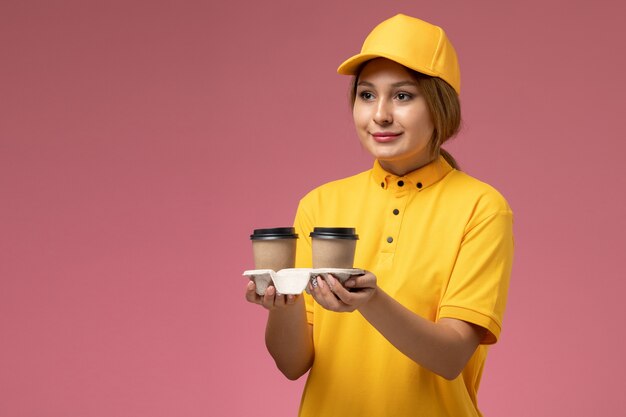 Front view female courier in yellow uniform yellow cape holding plastic brown coffee cups and delivering them on the pink desk uniform delivery female color