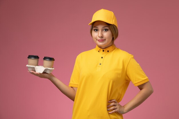 ピンクの背景にプラスチック製の茶色のコーヒーカップを保持している黄色の制服黄色のケープの正面図女性宅配便均一配達女性の色