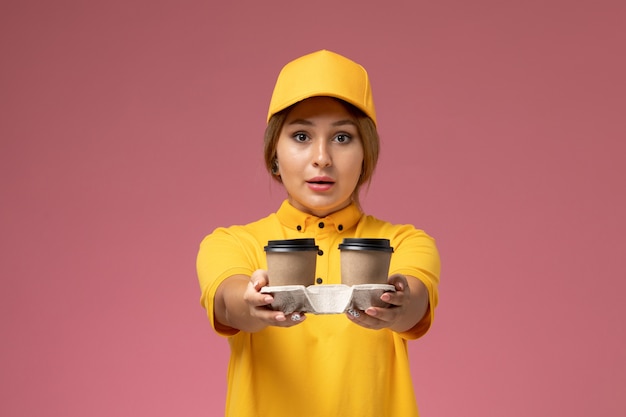 Вид спереди женщина-курьер в желтой униформе с желтым плащом держит пластиковые коричневые кофейные чашки на розовом столе.