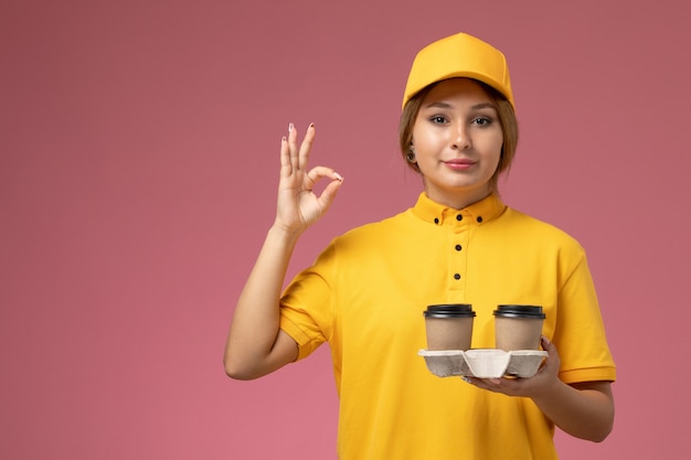 ピンクの机の上に大丈夫なサインを示すプラスチック製の茶色のコーヒーカップを保持している黄色の制服黄色のケープの正面図女性宅配便