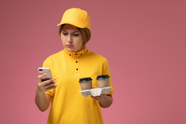 Front view female courier in yellow uniform yellow cape holding plastic brown coffee cups using a phone on the pink desk uniform delivery female