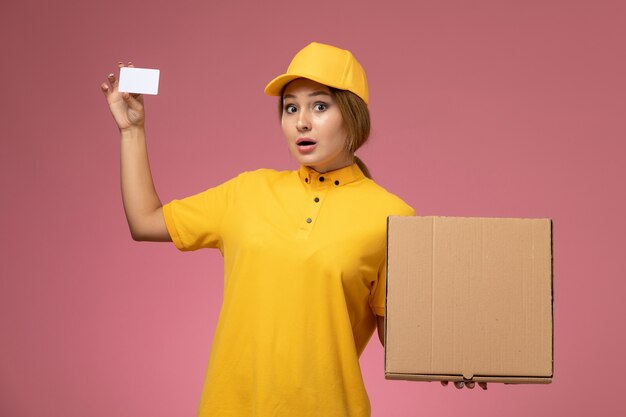 Front view female courier in yellow uniform yellow cape holding plastic card and food box on the pink desk uniform delivery female color