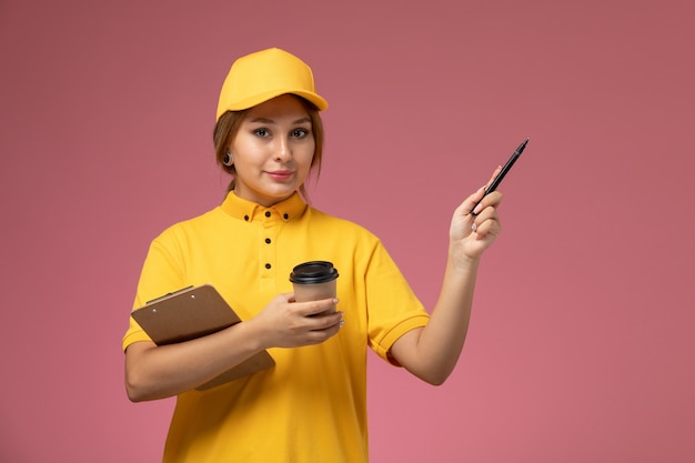 Вид спереди женщина-курьер в желтой униформе, желтой накидке, держащей пластиковую ручку для блокнота с чашкой кофе на розовом фоне.