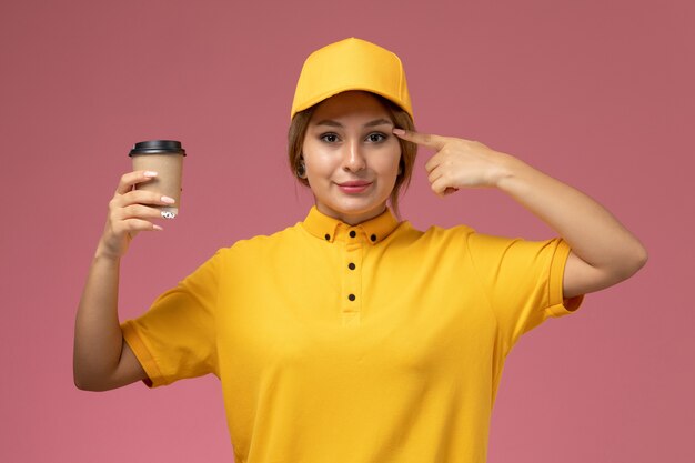 Free photo front view female courier in yellow uniform yellow cape holding plastic coffee cup on the pink background uniform delivery job work