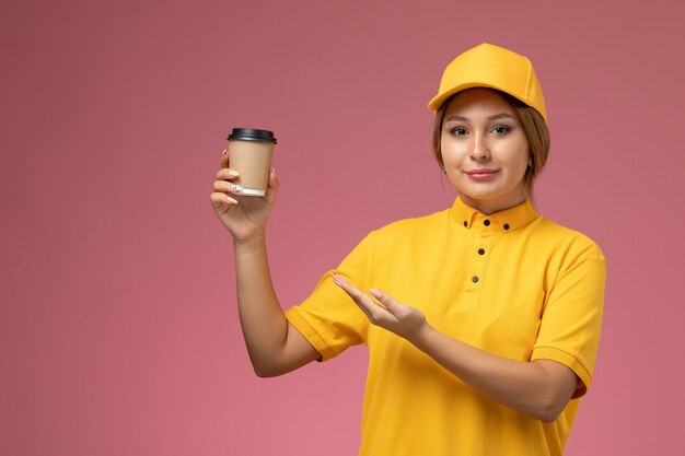 Вид спереди женщина-курьер в желтой форме с желтым плащом держит пластиковую кофейную чашку на розовом фоне.