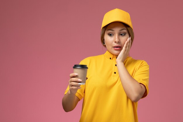 Front view female courier in yellow uniform yellow cape holding plastic coffee cup on the pink background uniform delivery work job