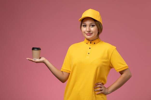 ピンクの背景に笑顔でプラスチック製のコーヒーカップを保持している黄色の制服黄色のケープの正面図女性宅配便制服配達作業の仕事