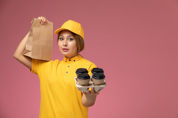 Foto gratuita corriere femminile di vista frontale in capo giallo uniforme giallo che tiene le tazze di caffè di plastica e il pacchetto di cibo sul lavoro di colore del lavoro di consegna uniforme del pavimento rosa