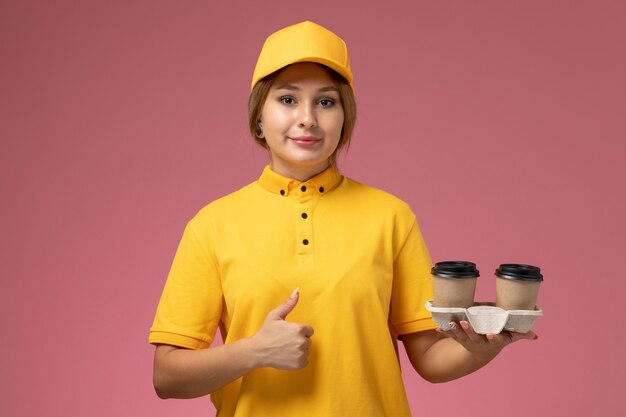 Front view female courier in yellow uniform yellow cape holding plastic coffee cups on pink background uniform delivery work job