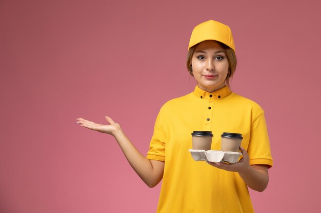 ピンクの背景にプラスチック製のコーヒーカップを保持している黄色の制服黄色のケープの正面図女性宅配便制服配達作業の仕事