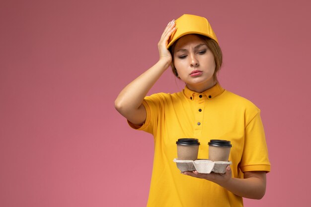 ピンクの背景にプラスチック製のコーヒーカップを保持している黄色の制服黄色のケープの正面図女性宅配便制服配達作業