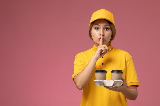 Front view female courier in yellow uniform yellow cape holding plastic coffee cups on pink background uniform delivery work