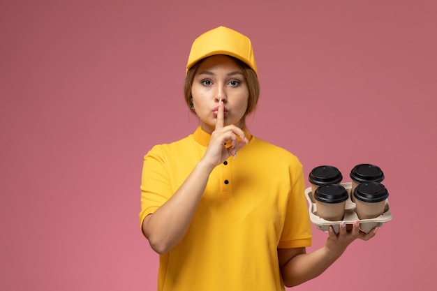 플라스틱 커피 컵을 들고 분홍색 배경 유니폼 배달 작업 색상 작업에 침묵 기호를 보여주는 노란색 유니폼 노란색 케이프 전면보기 여성 택배