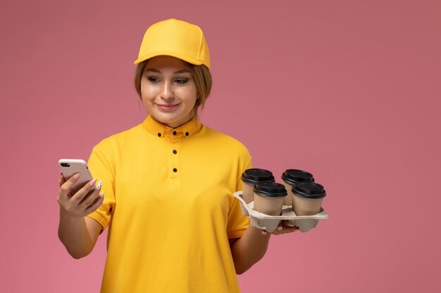 Front view female courier in yellow uniform yellow cape holding plastic coffee cups and using phone on pink desk uniform delivery color job