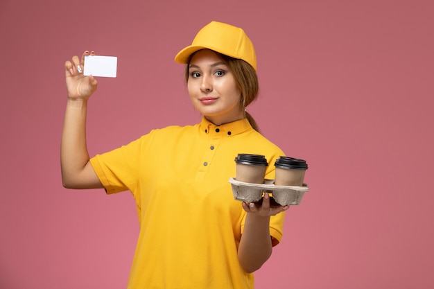 Front view female courier in yellow uniform yellow cape holding plastic coffee cups white card on the pink desk uniform delivery work color job