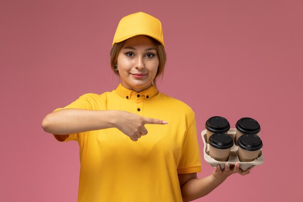 Front view female courier in yellow uniform yellow cape holding plastic coffee cups with a slight smile on the phone on the pink desk uniform delivery work color job