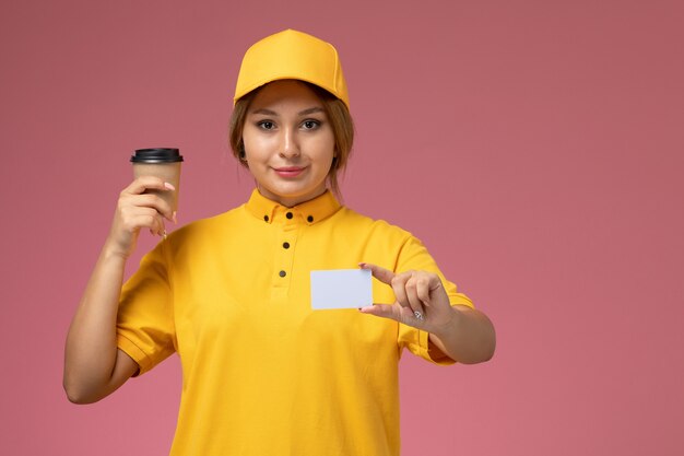 ピンクの背景の制服配達の仕事で白いカードとコーヒーを保持している黄色の制服黄色の岬の正面図女性宅配便