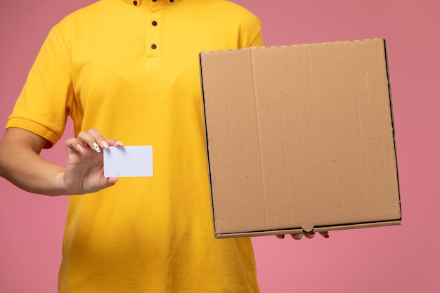 Front view female courier in yellow uniform yellow cape holding white card with food box on the pink desk uniform delivery female color