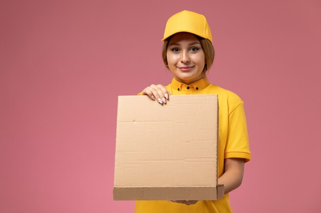 Front view female courier in yellow uniform yellow cape opening food box and smiling on the pink desk uniform delivery female color