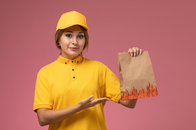 Front view female courier in yellow uniform yellow cape smiling holding food package on pink background uniform delivery work color job