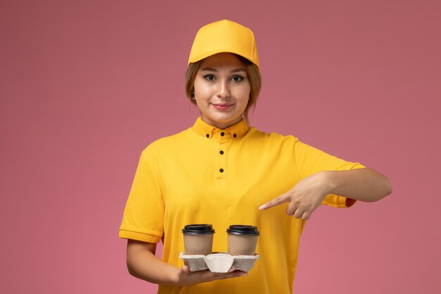 ピンクの机の上のプラスチック製の茶色のコーヒーカップを保持している黄色の制服黄色のケープ笑顔の正面図女性宅配便制服配達女性の色