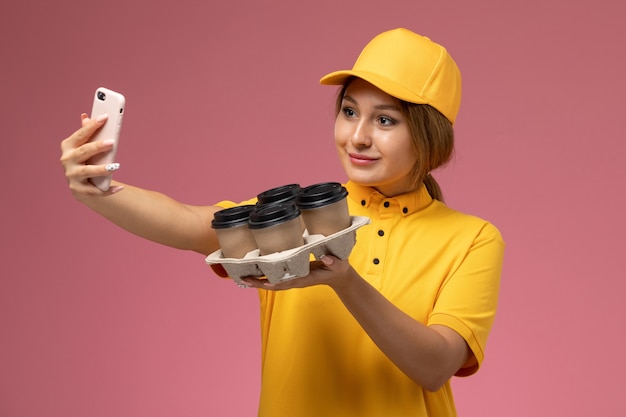 ピンクの背景にコーヒーカップとselfieを取っている黄色の制服黄色のケープの正面図女性宅配便制服配達作業色の仕事