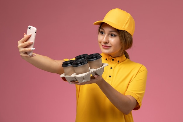 Front view female courier in yellow uniform yellow cape taking a selfie with coffee cups on pink background uniform delivery work color job