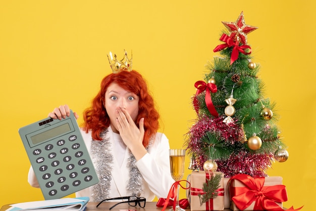 Вид спереди женщина-врач, держащая калькулятор вокруг рождественских подарков и дерева