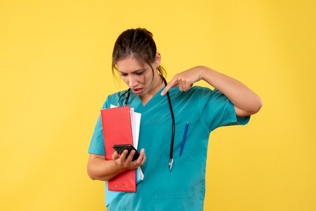 黄色の背景に分析と電話を保持している医療シャツの正面図の女性医師