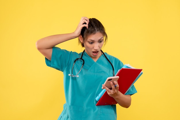 Front view female doctor in medical shirt holding analysis and phone on yellow background