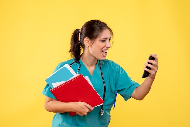 黄色の背景に分析と電話を保持している医療シャツの正面図の女性医師
