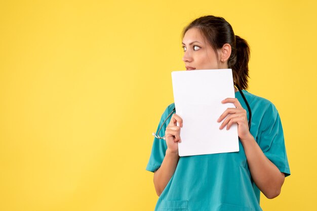 黄色の背景に紙の分析を保持している医療シャツの正面の女性医師