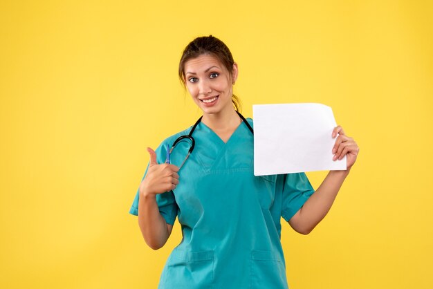 黄色の背景に紙の分析を保持している医療シャツの正面の女性医師